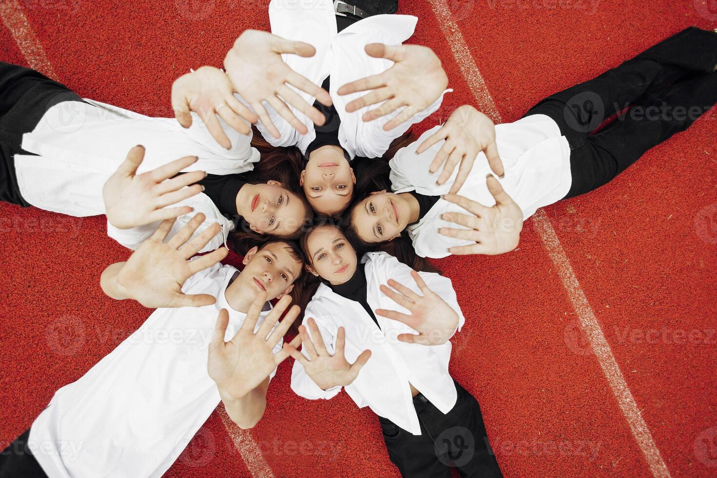 Happy friends lying together in a circle looking at the camera, holding hands and smiling while lying on a red wall... Friendship, lifestyle, togetherness, business and teamwork concepts. photo