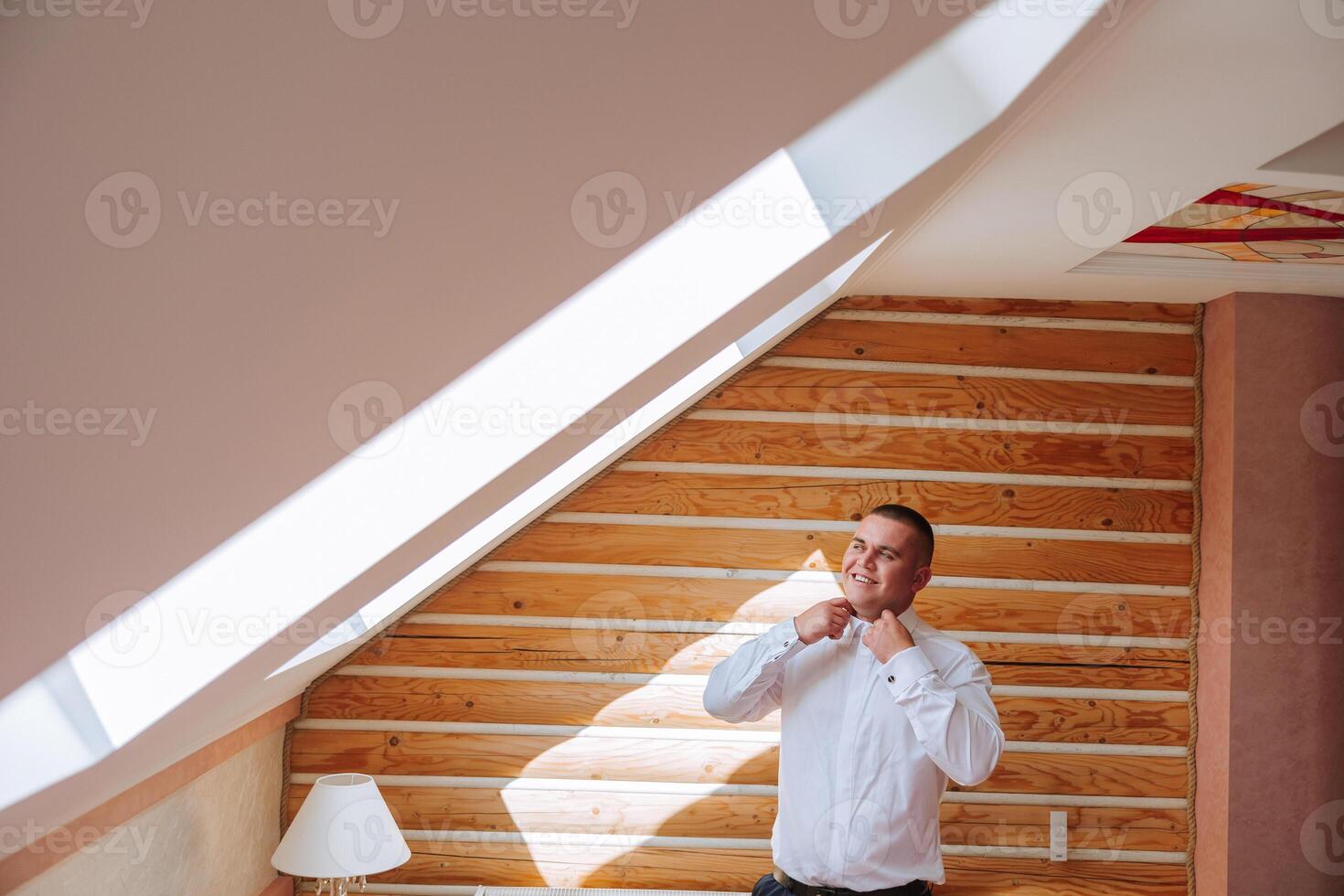 The groom buttons his shirt by the window. man in a shirt. Male portrait. Photoshoot at the window. Groom's collection. Wedding day. Handsome man groom buttoning his shirt in a hotel room. photo