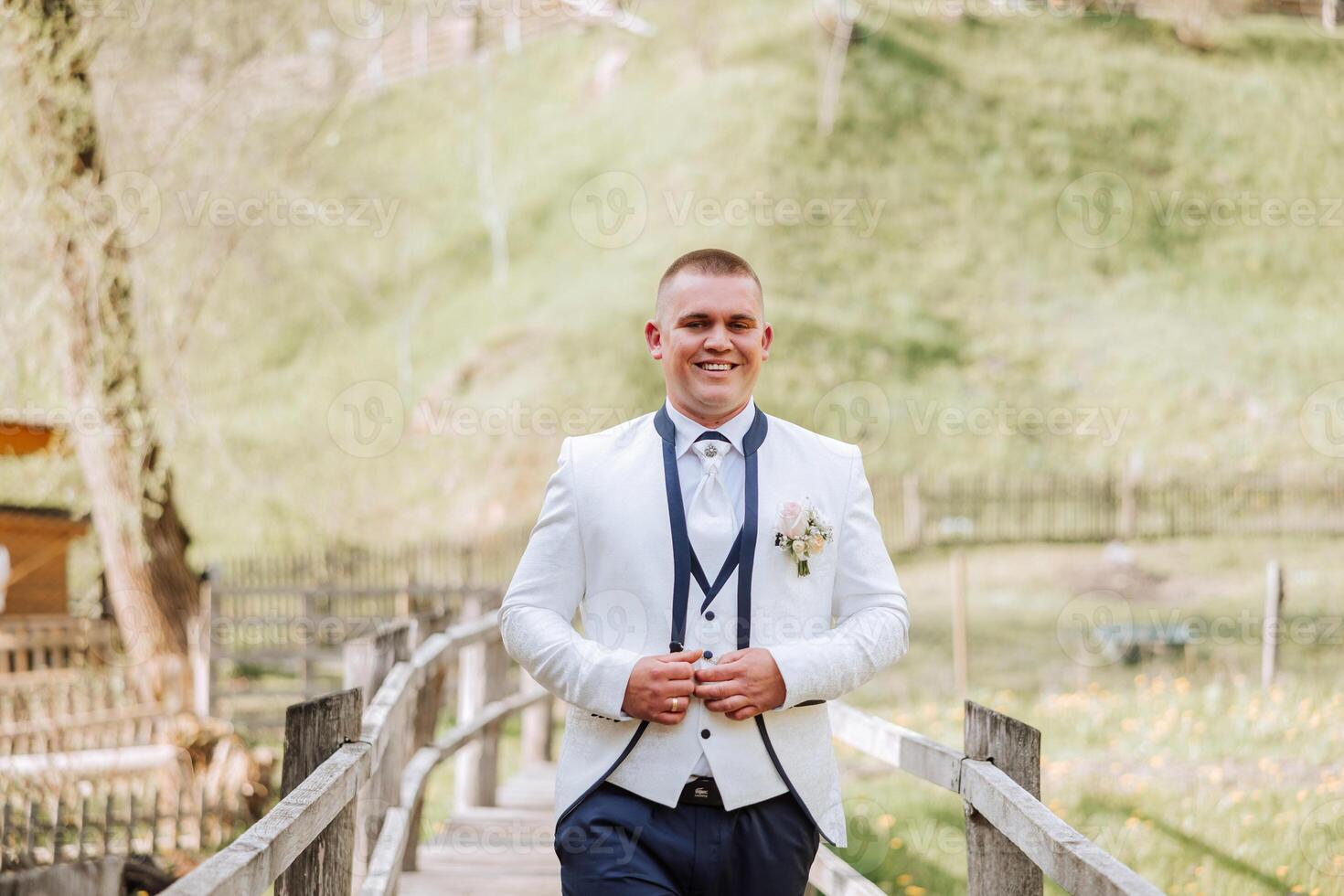 The groom in a white jacket and black pants poses smiling and fastens the button on his shirt. Wedding portrait. photo