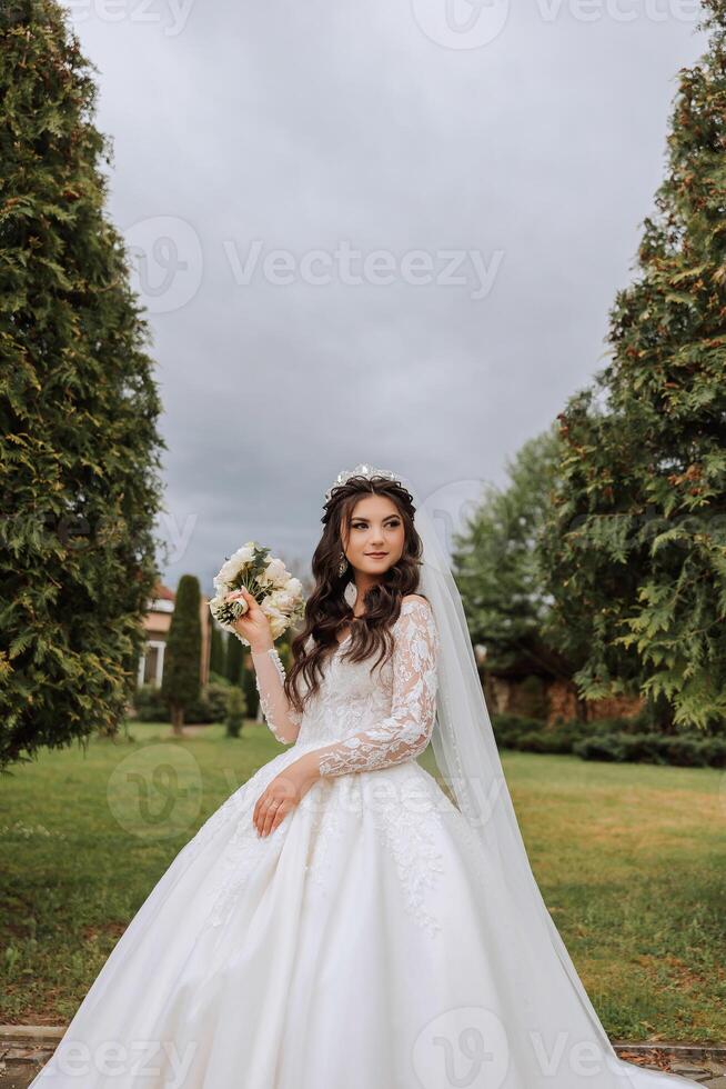 Rizado morena novia en un lozano velo y vestir con largo mangas, participación un ramo de flores y posando en contra un antecedentes de verde arboles primavera Boda foto