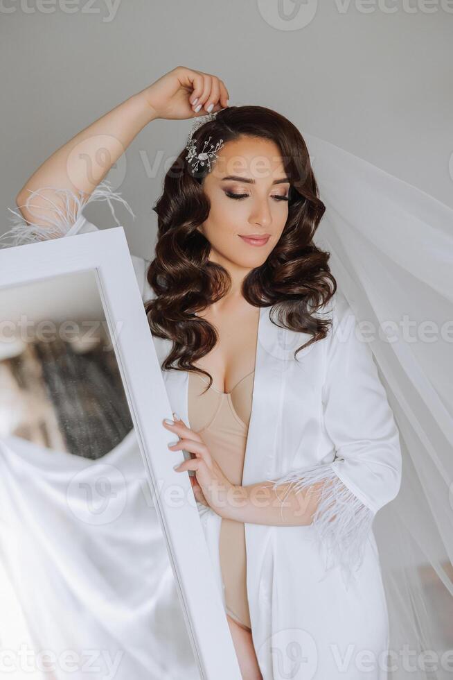 portrait of a luxurious bride in boudoir style near a mirror with reflection. Preparing the bride for the wedding in the morning in a white robe. photo