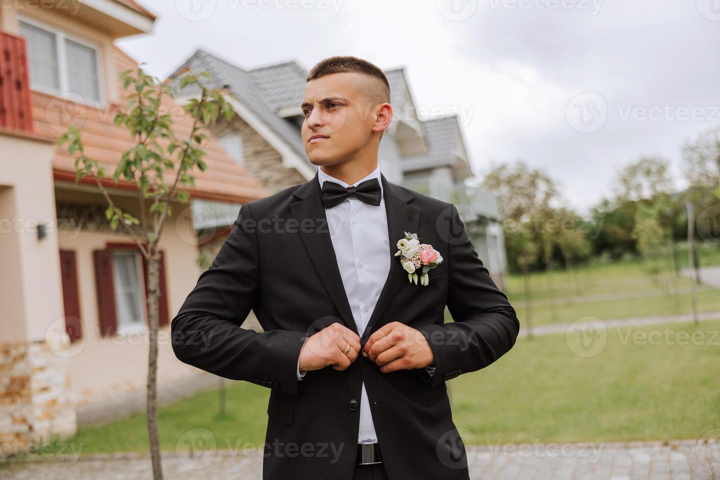 The groom in a black suit adjusts his jacket, poses against the background of a green tree. Wedding portrait. photo