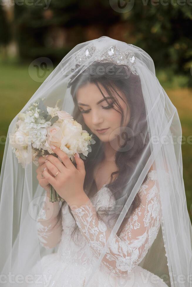 el novia en un lujoso vestido, envuelto en un velo, olfatea su ramo de flores y poses para el cámara. magnífico vestir con largo mangas, abierto busto. primavera Boda foto