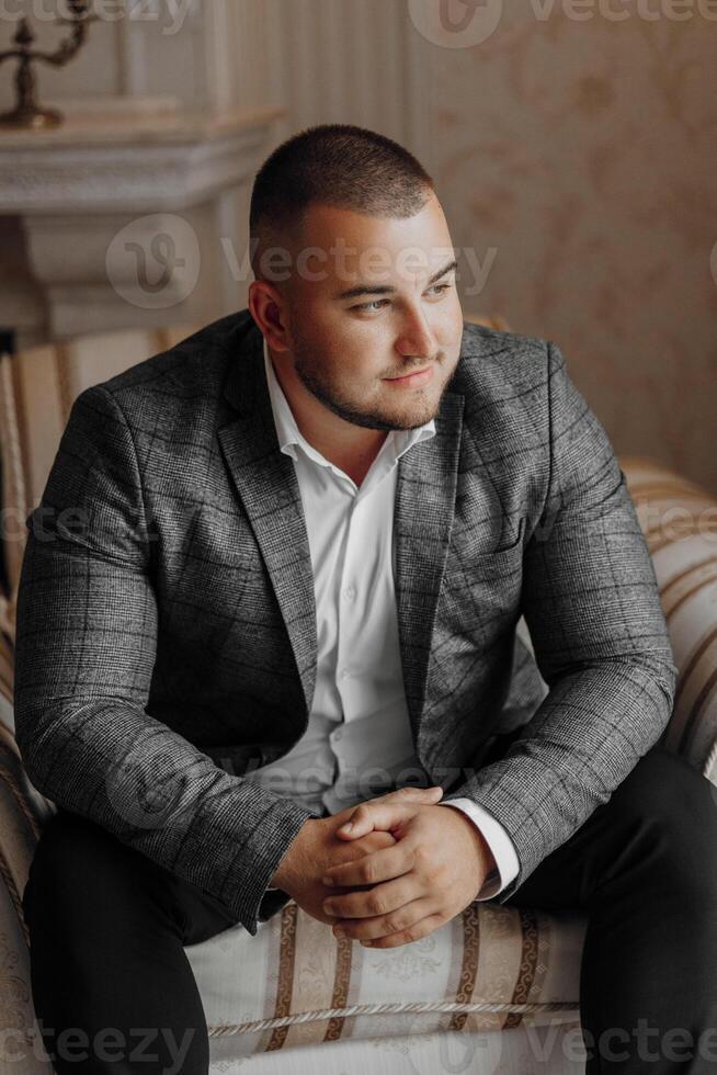 a handsome and positive man is sitting on an expensive chair. Smiling at the camera, wearing smart shoes, trousers, a jacket and a white shirt photo