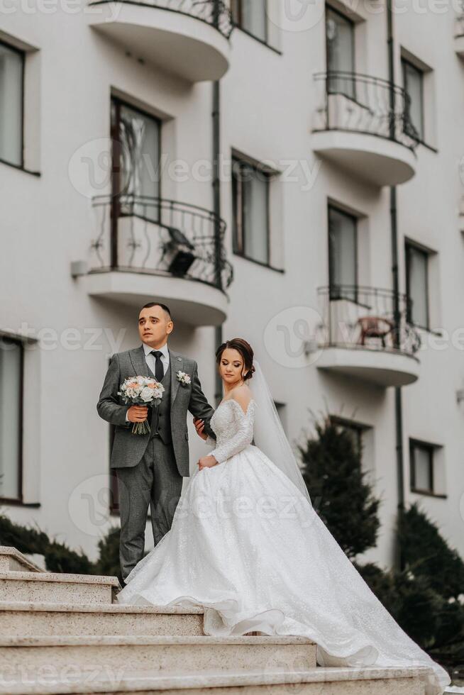 A bride in a white dress with a train and a groom in a suit pose on the steps of a building. Wedding photo session in nature