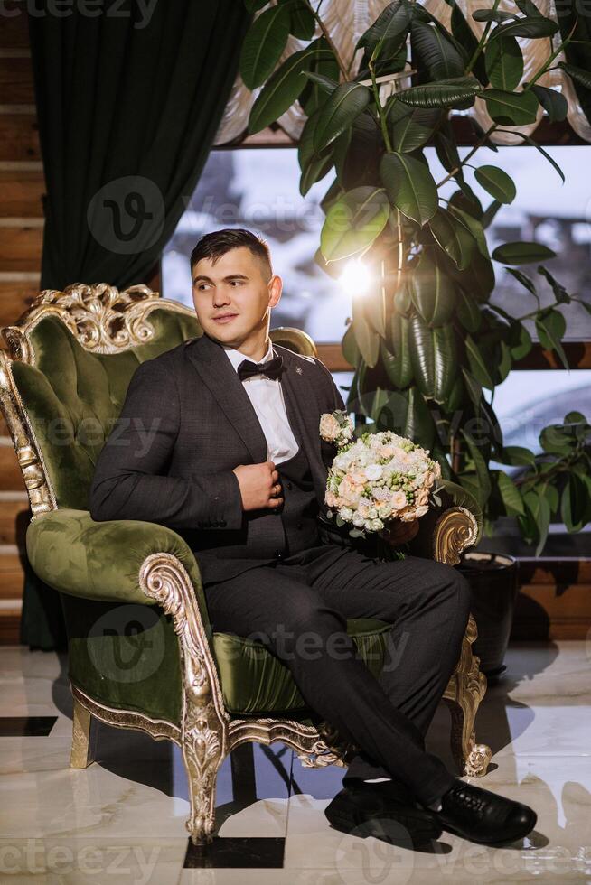 A stylish groom sits on a chair in front of a large window. A man is holding a wedding bouquet of flowers. Wedding day. photo