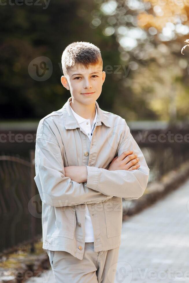 Vertical close-up portrait of a teenager in casual clothes. Happy smiling teenager in autumn park in sunlight. A beautiful child looks at the camera in nature. photo