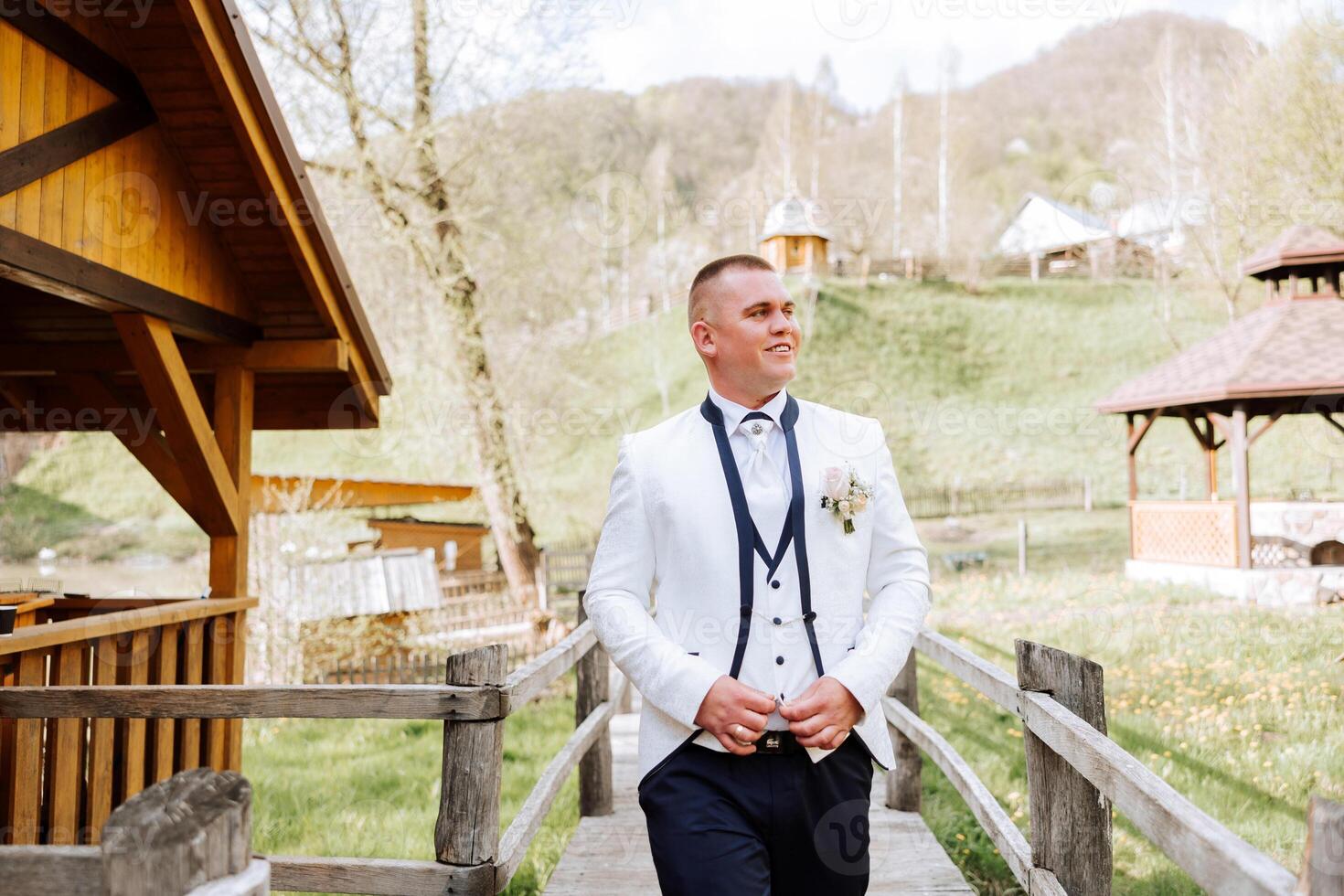 The groom in a white jacket and black pants poses smiling and fastens the button on his shirt. Wedding portrait. photo