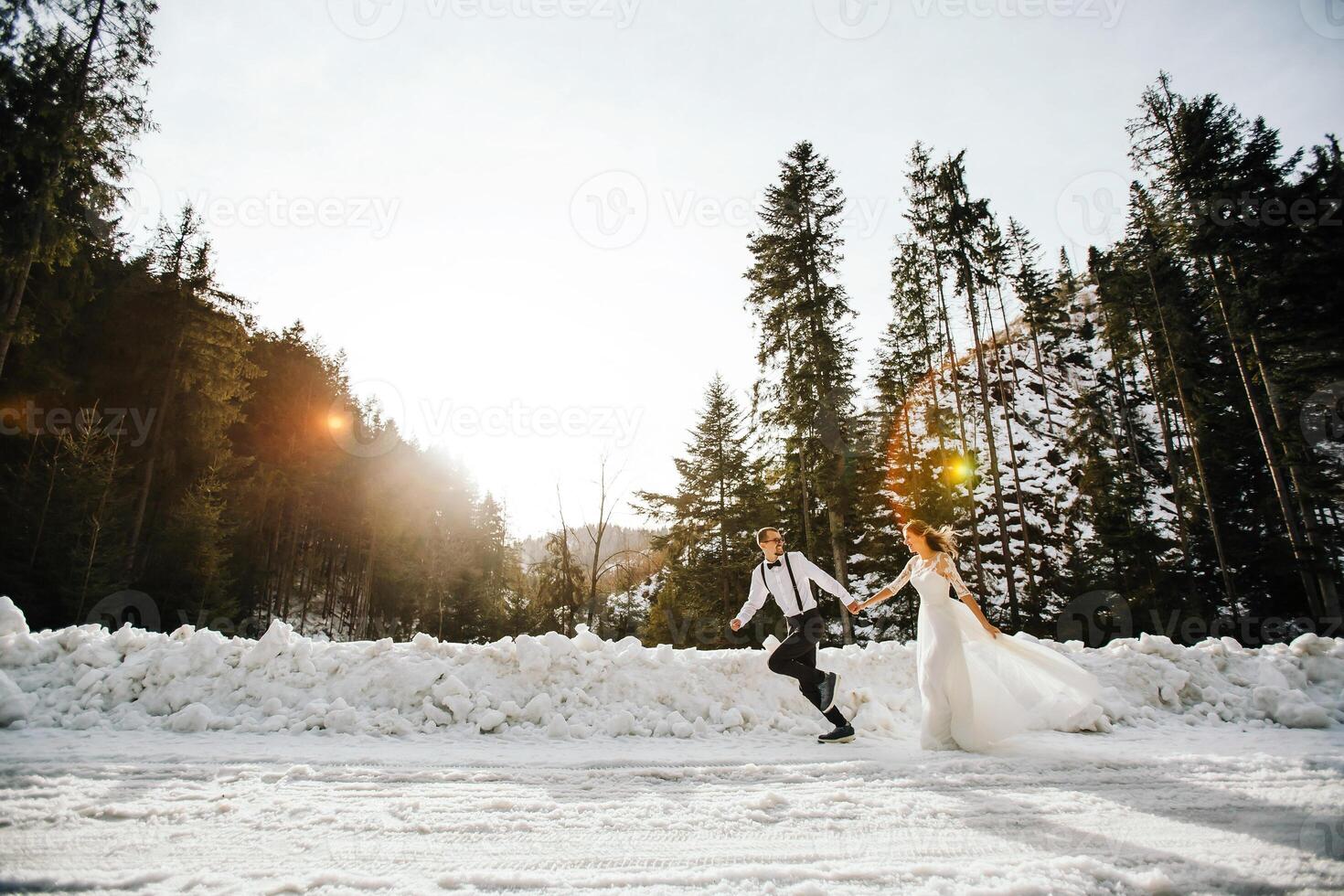 The bride and groom are running along a snowy road against the background of a pine forest and beautiful contrast sunlight. Side view. Winter wedding. Place for logo. photo