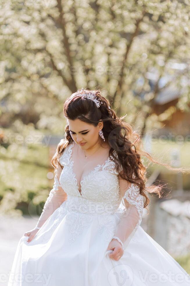 Curly brunette bride poses wrapped in a veil against the background of mountains and wooden houses. Magnificent dress with long sleeves, open bust. Summer wedding photo
