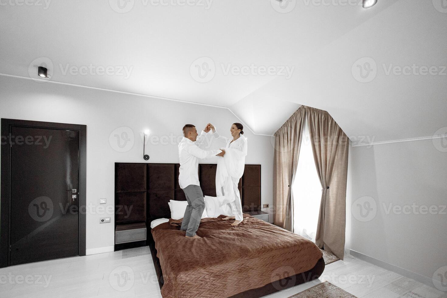 The morning of the bride and groom in a hotel room. Happy and in love bride and groom jumping and having fun on the bed. Confident girl-bride and handsome groom. Preparation for the wedding photo