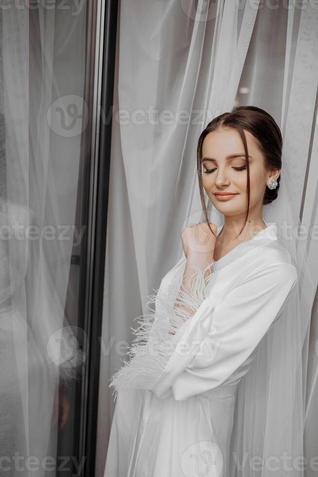 retrato de novia niña en blanco vestido con profesional peinado y natural maquillaje en hotel habitación con reflexión en ventana. el mejor día foto