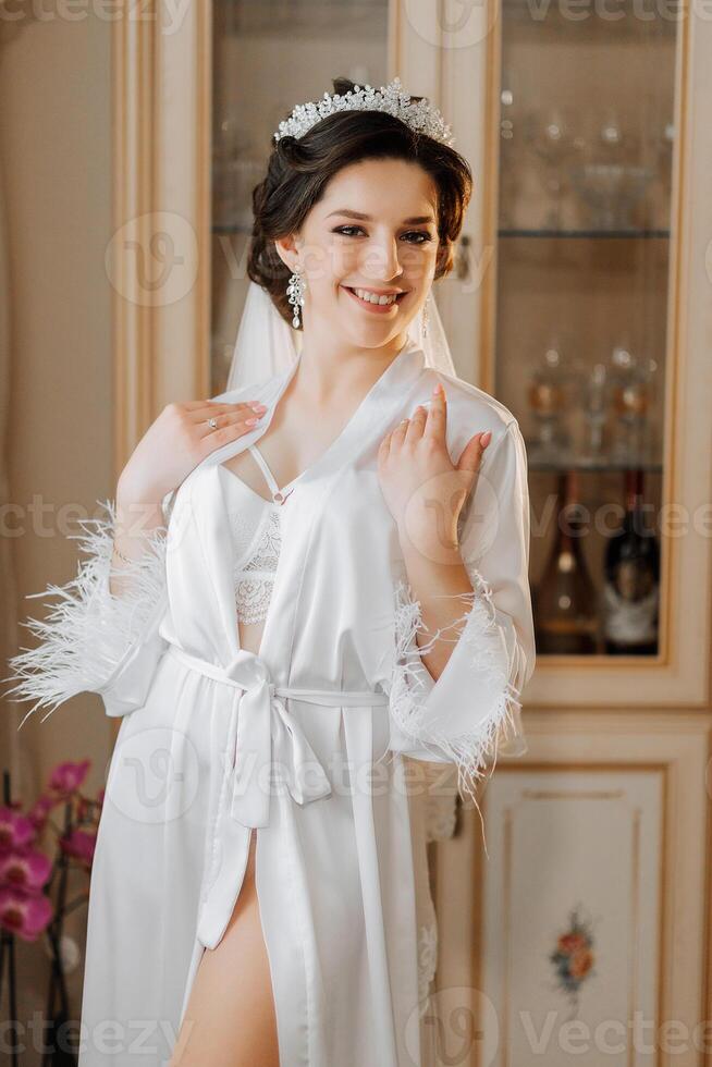 Morning of the bride before the wedding. A beautiful young woman with a veil and a beautiful hairstyle in a white robe in a room with a wonderful interior. Natural beauty and professional makeup. photo