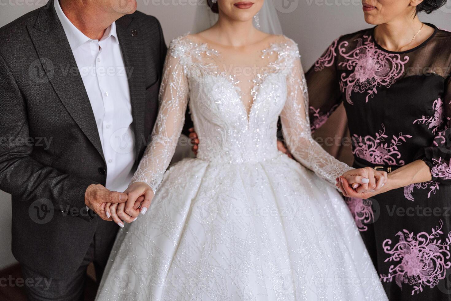 Happy parents hold the hands of the bride who is getting married and support her, wearing a wedding dress and a long veil. photo