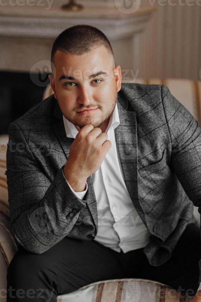 a handsome and positive man is sitting on an expensive chair. Smiling at the camera, wearing smart shoes, trousers, a jacket and a white shirt photo