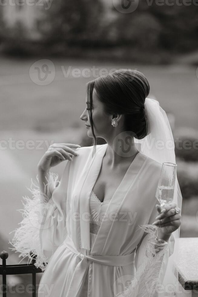 un hermosa novia con un largo velo en su cabello, vistiendo un elegante blanco bata, en pie en un balcón con vista a el ciudad, Bebiendo un bebida durante el Boda Mañana. negro y blanco foto