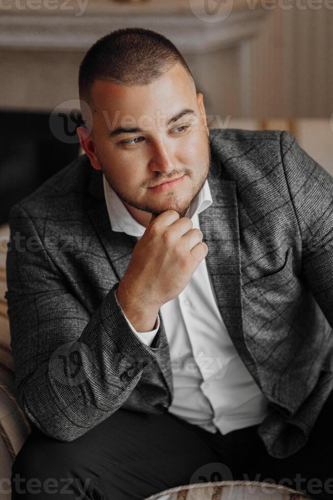 a handsome and positive man is sitting on an expensive chair. Smiling at the camera, wearing smart shoes, trousers, a jacket and a white shirt photo