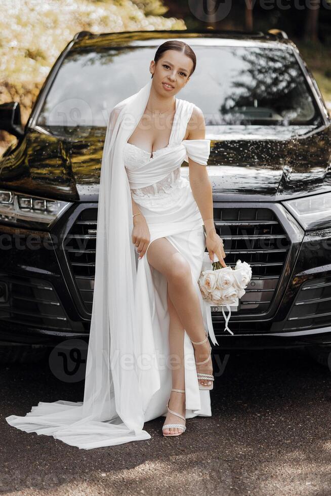 A dark-haired bride stands next to a modern black car on your wedding day with a bouquet of flowers. Portrait of the bride. White dress, long veil photo