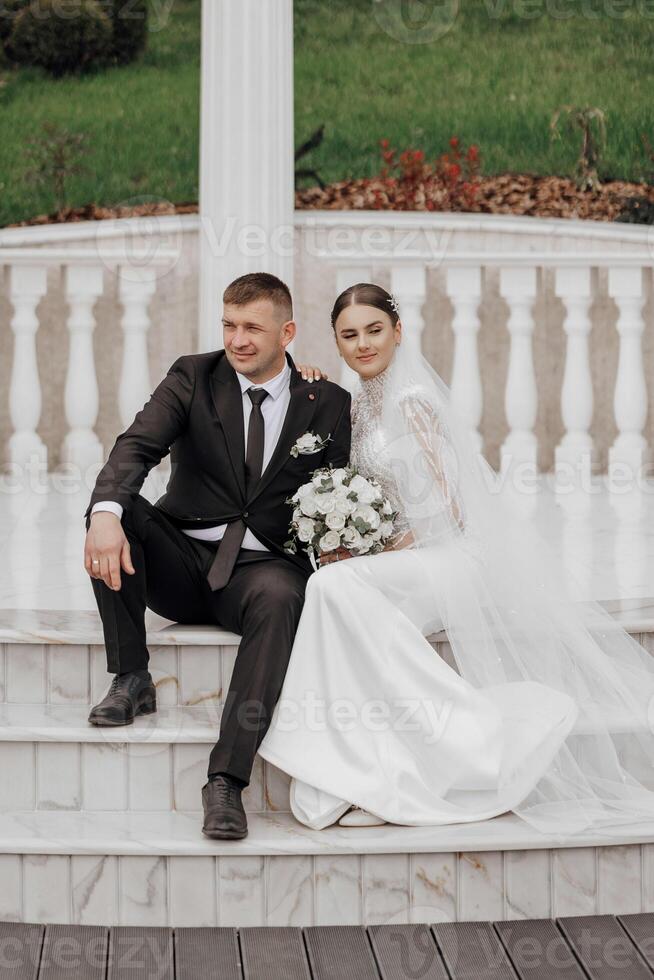 A bride in a long dress and a groom in a black suit sit near white Roman-style columns. Beautiful hair and makeup. An exquisite wedding photo