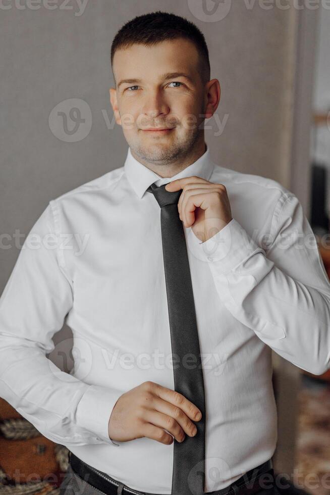 A man in a white shirt poses and adjusts his black tie. Portrait of the groom. Men's style. Business. Fashion photo