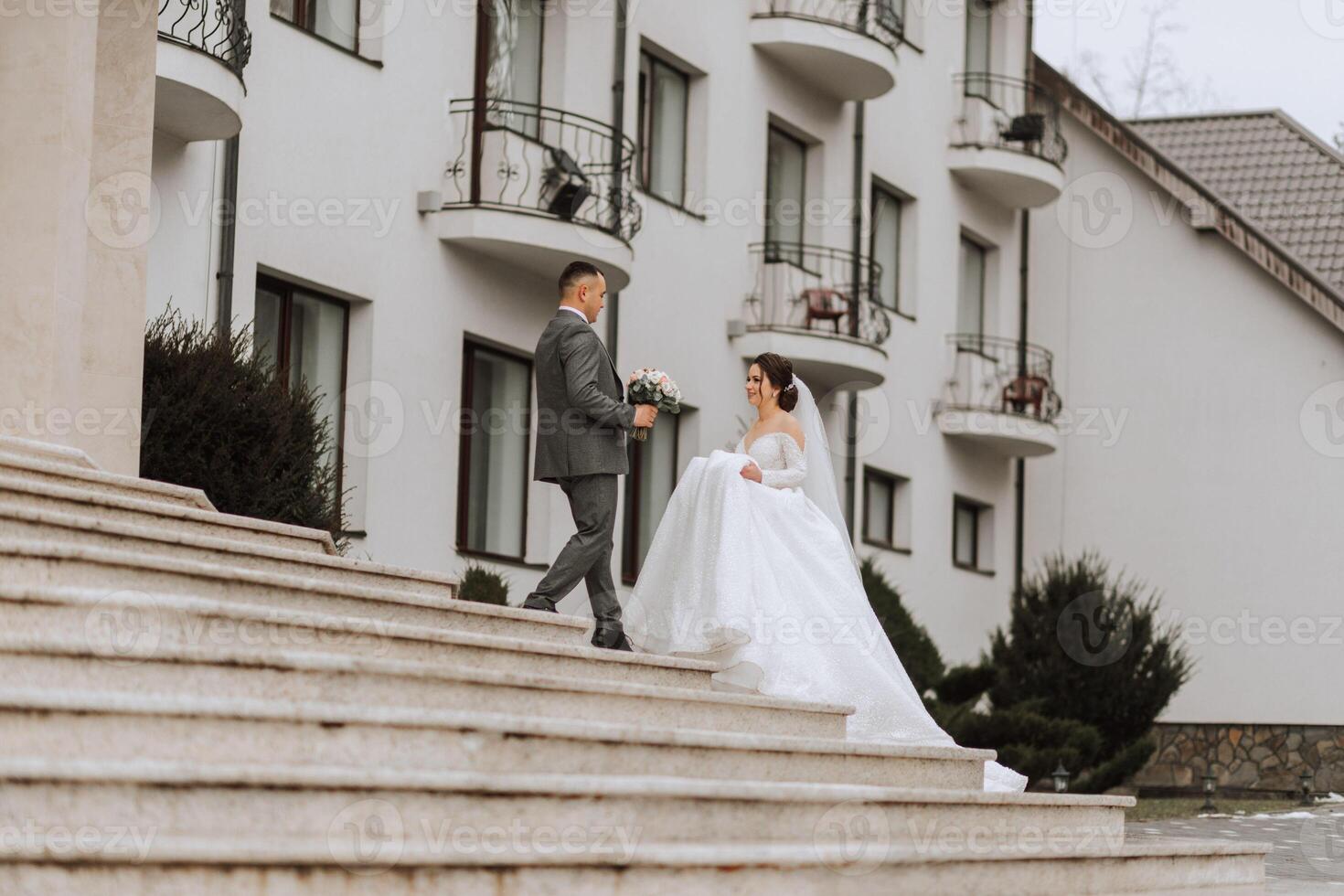 A bride in a white dress with a train and a groom in a suit pose on the steps of a building. Wedding photo session in nature
