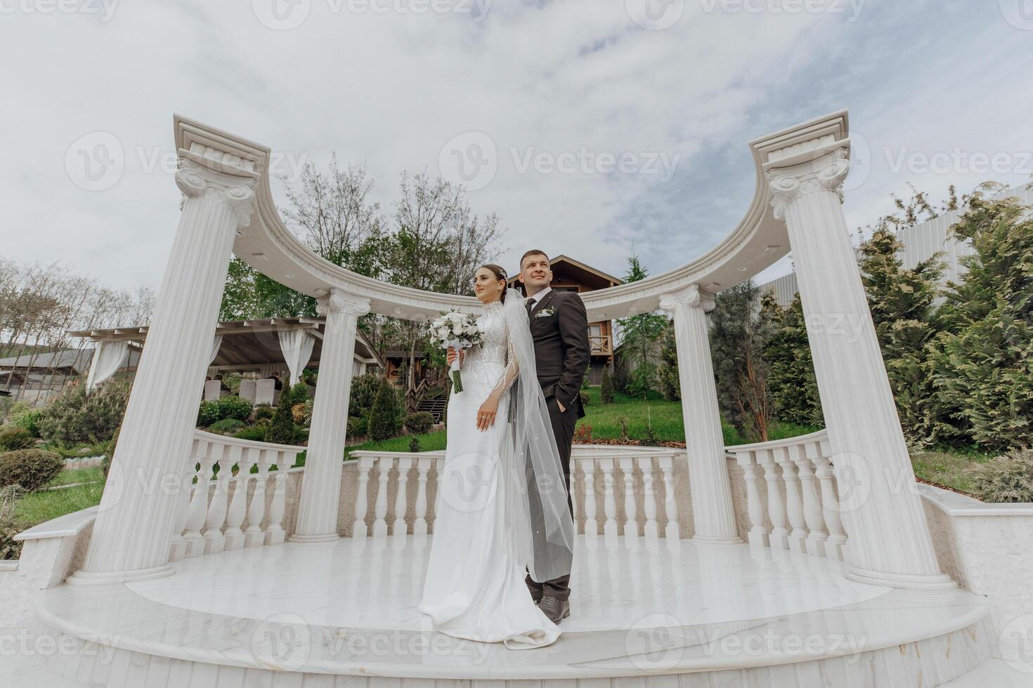 un novia en un largo vestir y un novio en un negro traje son en pie cerca blanco estilo romano columnas hermosa pelo y constituir. un Exquisito Boda foto