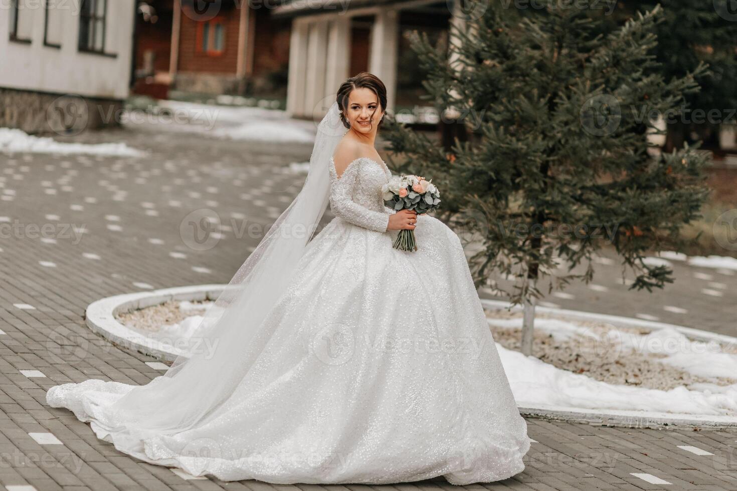 The bride in a white dress with a train and a long veil poses in nature, holding a bouquet. Walk. Winter wedding in nature. photo