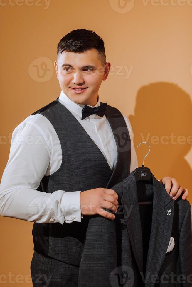 a young smiling man in a white shirt wears a bow tie and a black waistcoat. A groom with a short hairstyle on an orange background puts on his black suit and prepares for the wedding photo