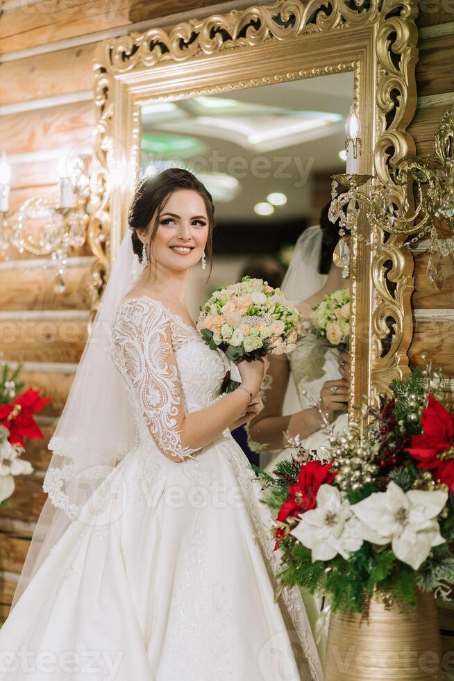 elegante morena novia poses en un blanco vestir cerca el espejo. retrato de el novia, Boda maquillaje y peinado, Boda moda. hermosa novia en un velo. foto