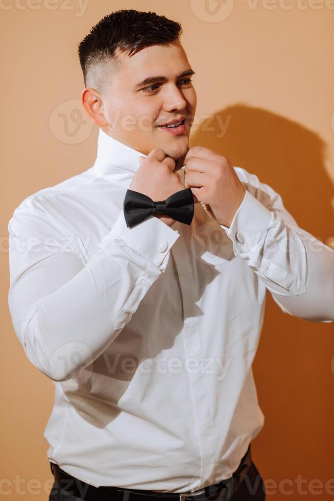 a young smiling man in a white shirt puts on a bow tie. The groom with a short hairstyle on an orange background is preparing for the wedding photo