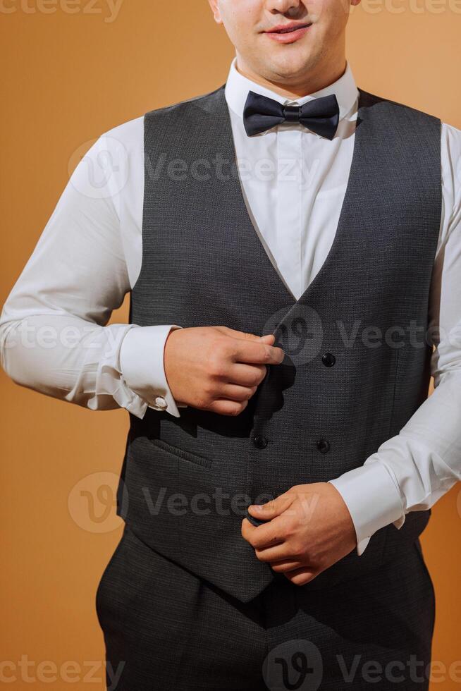 a young smiling man in a white shirt wears a bow tie and a black waistcoat. A groom with a short hairstyle on an orange background puts on his black suit and prepares for the wedding photo