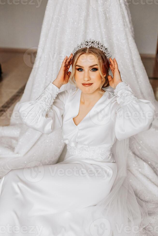 A bride in a silk suit with a veil poses in her room, sitting on the floor next to her wedding dress. Morning of the bride. Preparation photo