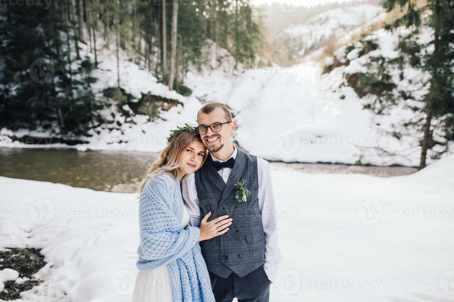 Bride and groom on the background of a pine forest and a river. The bride in a white wedding dress is wrapped in a blue blanket, the groom is in a white shirt and waistcoat. Winter wedding. photo