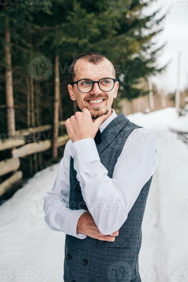 Boda retrato de el novio. el novio soportes en contra el antecedentes de el invierno bosque. un hombre en un chaleco y blanco camisa, lentes y un arco atar. invierno Boda foto