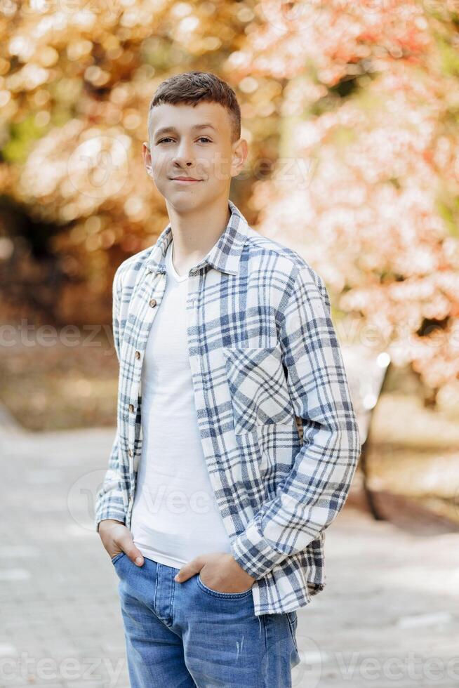 Vertical close-up portrait of a teenager in casual clothes. Happy smiling teenager in autumn park in sunlight. A beautiful child looks at the camera in nature. photo