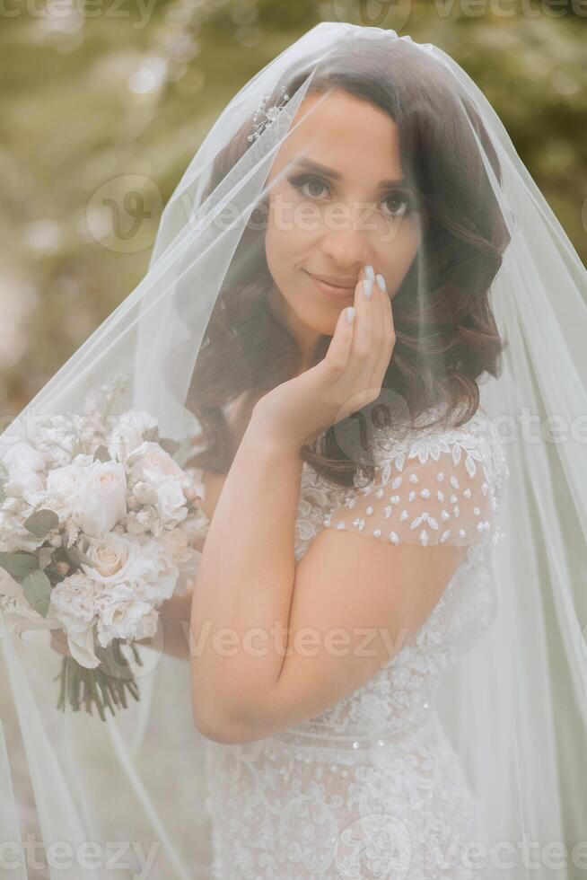 A beautiful bride is standing in a green spring forest in a glamorous white wedding dress under a veil. A romantic beautiful bride in a white dress is posing against the background of the forest. photo