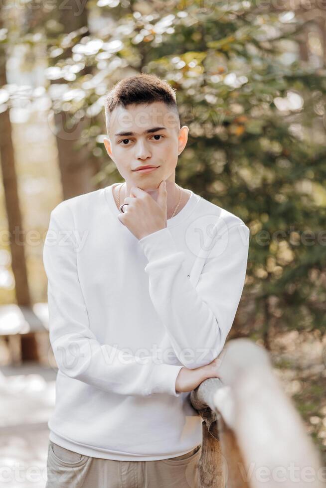 Close-up vertical portrait of a teenager in a white sweater and brown pants. Happy smiling teenager in summer park in sunlight. A beautiful child is looking at the camera in the clearing. photo