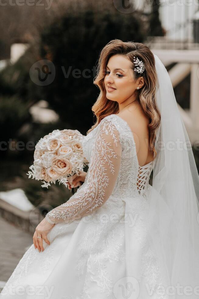 Portrait of a beautiful bride with a wedding bouquet of flowers, attractive woman in a wedding dress with a long veil. Happy bride woman. Bride with wedding makeup and hairdo. Winter wedding photo