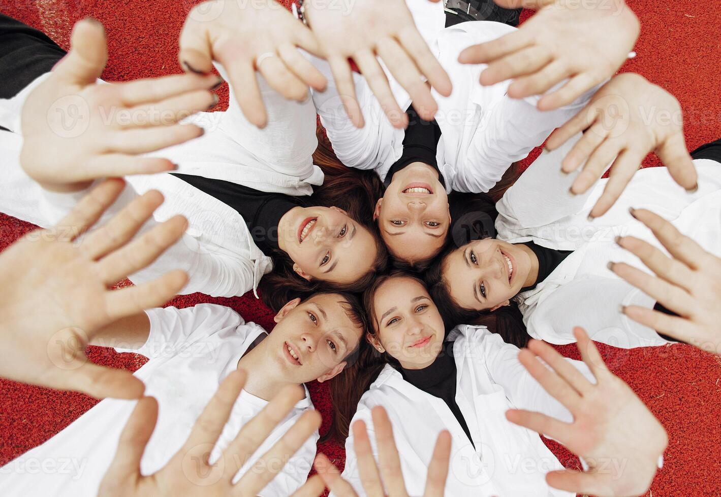 Happy friends lying together in a circle looking at the camera, holding hands and smiling while lying on a red wall... Friendship, lifestyle, togetherness, business and teamwork concepts. photo