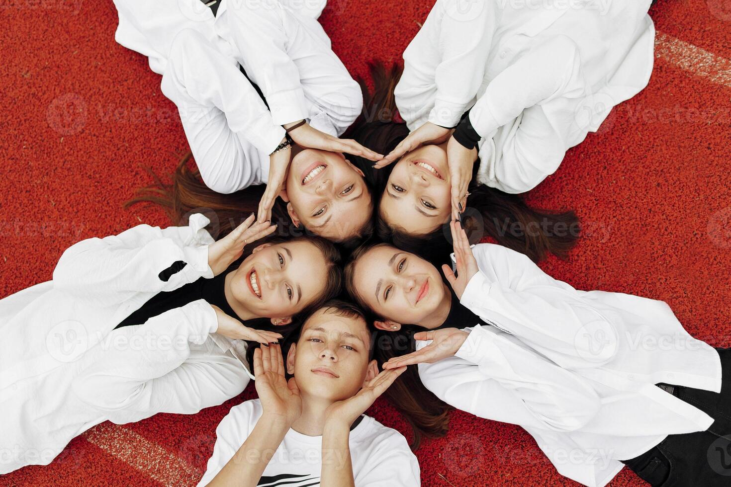 Happy friends lying together in a circle looking at the camera, holding hands and smiling while lying on a red wall... Friendship, lifestyle, togetherness, business and teamwork concepts. photo