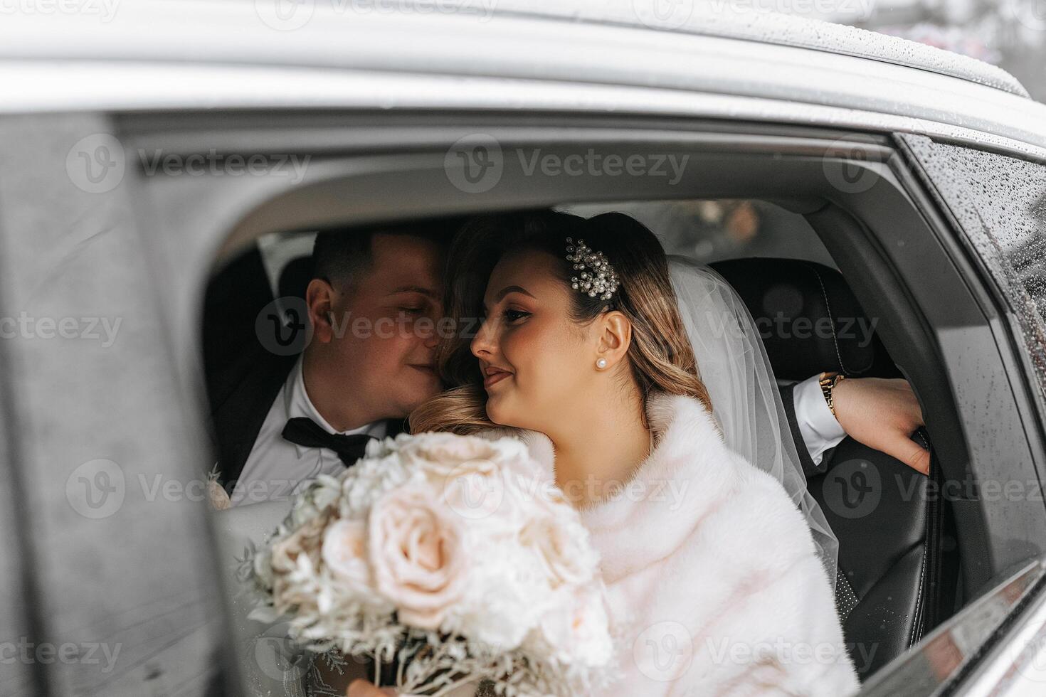 Portrait of incredibly beautiful brides in the window of an expensive car. The bride with a bouquet of flowers looks at her groom. photo