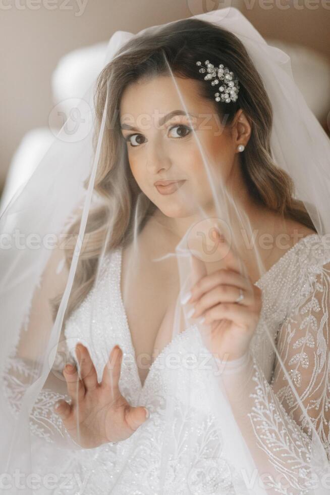 retrato de un oferta novia en un Boda vestir y un hermosa peinado debajo un velo en su habitación desde el Mañana foto