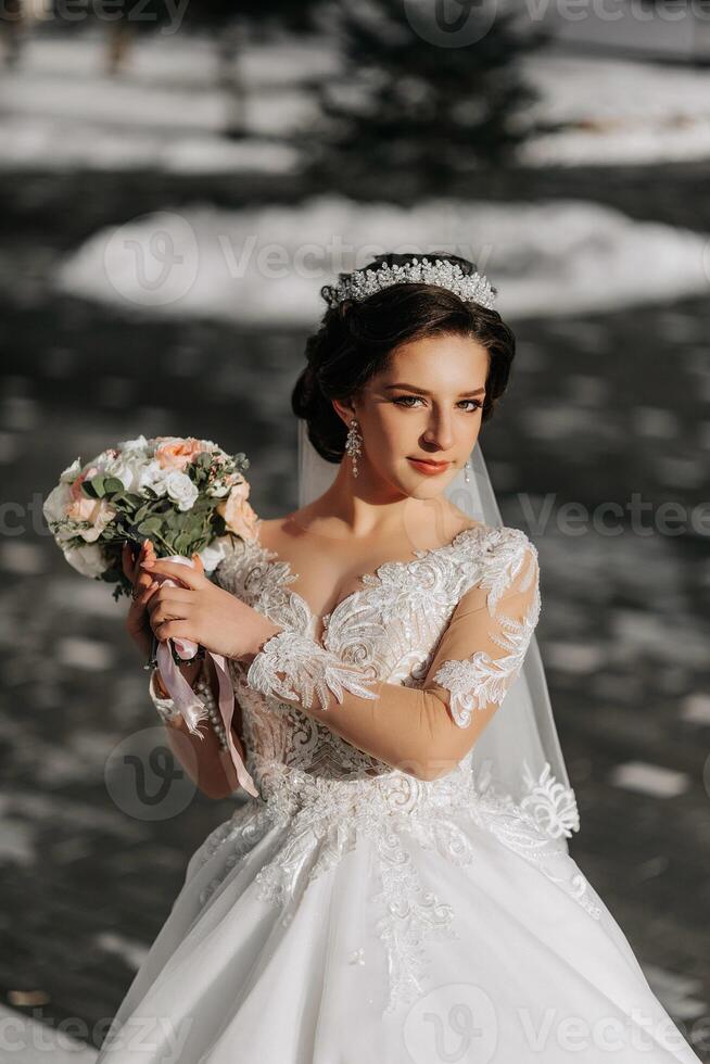 Portrait of a beautiful bride with a wedding bouquet of flowers, attractive woman in a wedding dress with a long veil. Happy bride woman. Bride with wedding makeup and hairdo. Winter wedding photo