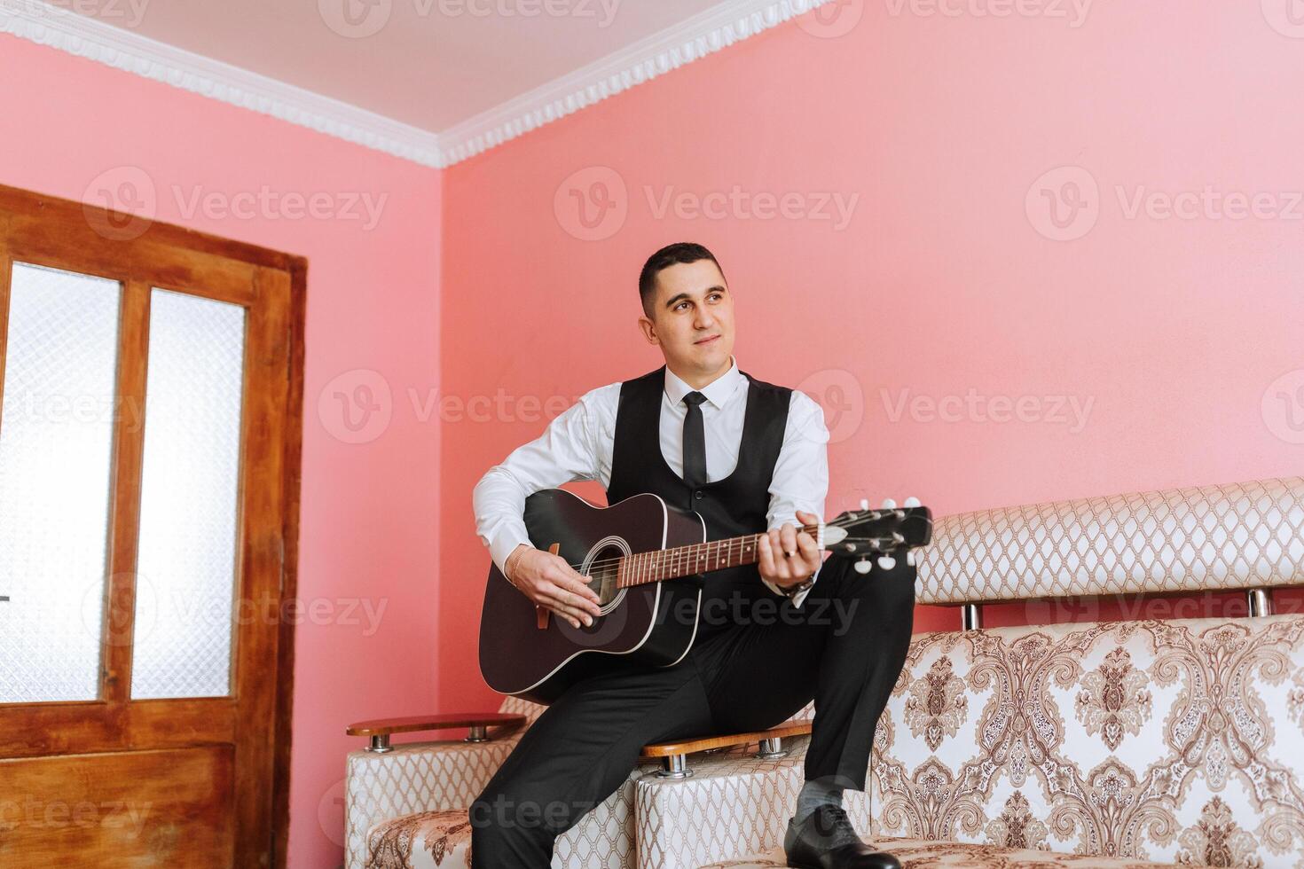 The groom sings songs to the guitar in the morning in his room. Groom in a black suit. Happy man. Preparation for the wedding. photo