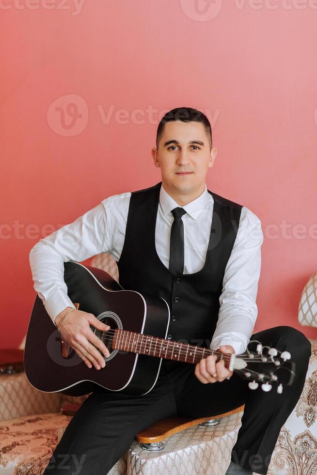 The groom sings songs to the guitar in the morning in his room. Groom in a black suit. Happy man. Preparation for the wedding. photo