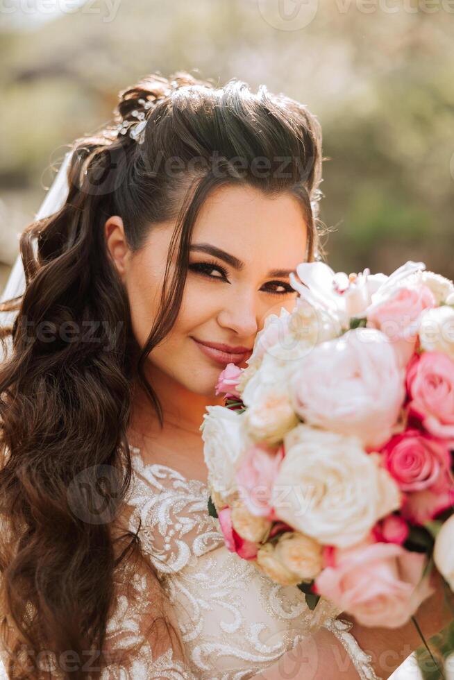 Rizado morena novia poses con un ramo, cerca un de madera barandilla. magnífico vestir con largo mangas, abierto busto. primavera Boda foto