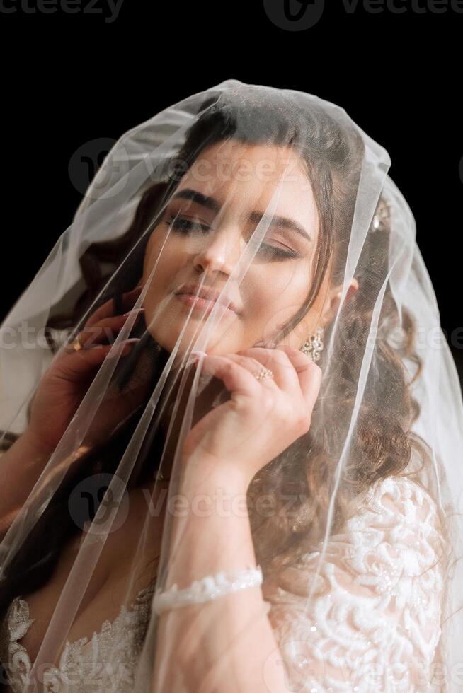 Curly brunette bride poses wrapped in a veil against the background of mountains and wooden houses. Magnificent dress with long sleeves, open bust. Summer wedding photo