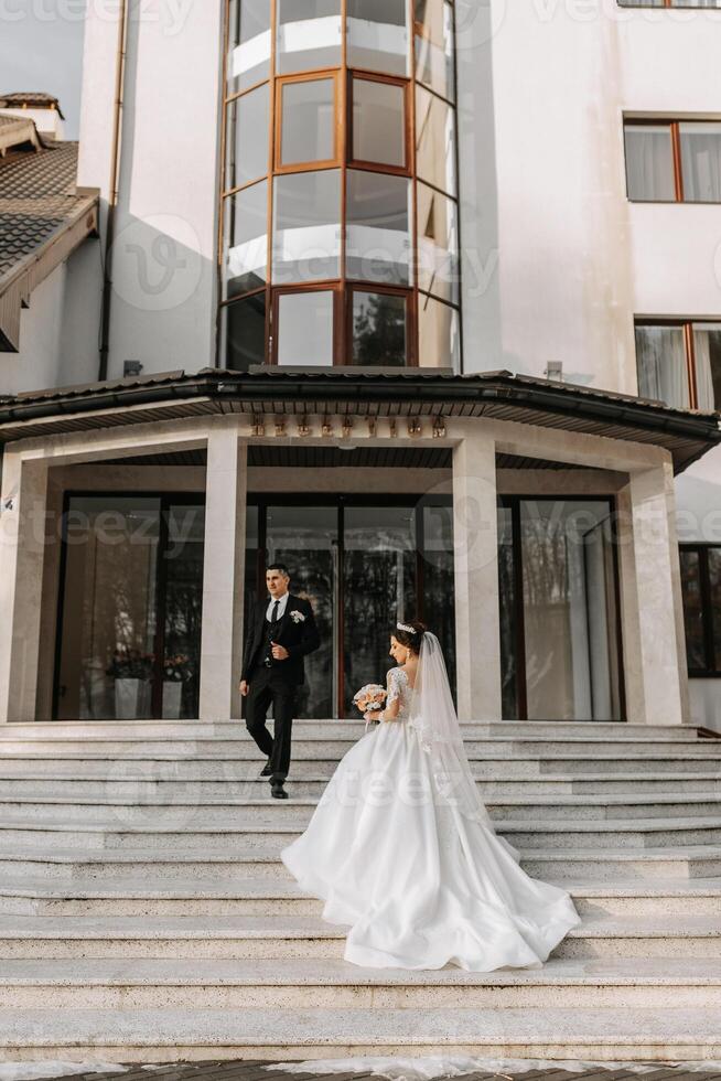 The bride and groom are walking near the hotel and posing, happy and enjoying the day, holding hands. A long train on the dress. Winter wedding photo