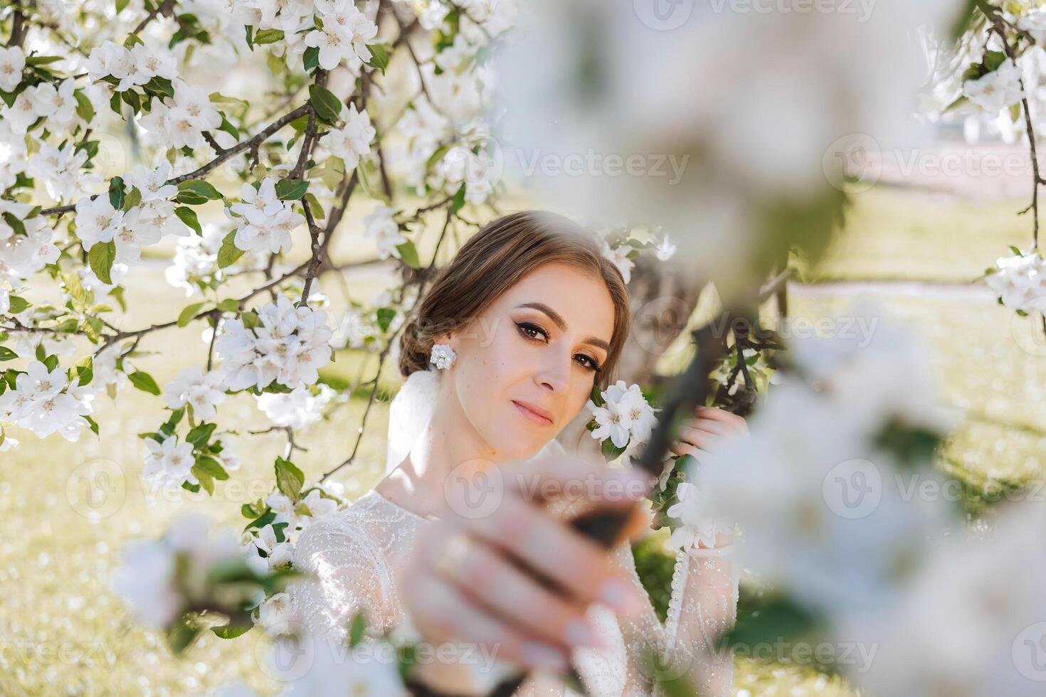 A red-haired bride poses against the background of a blooming tree. Magnificent dress with long sleeves, open bust. Spring wedding photo