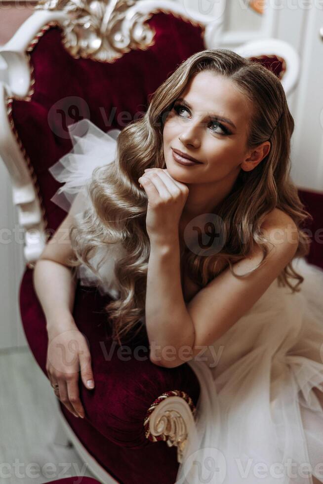 Portrait of a young beautiful elegant woman sitting and posing on a red suede armchair in Art Nouveau style. Beautiful model girl in a luxurious evening dress. Lady with perfect makeup and wavy hair. photo